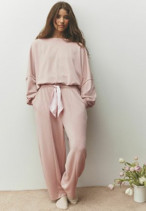 Junge Frau mit langen Haaren, die lockere rosa Loungewear trägt, steht barfuß an einer weißen Wand neben einer Vase mit rosa und weißen Blumen.