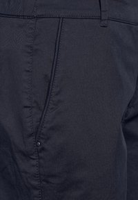 Close-up van een donkerblauwe broekzak met zichtbare stiksels en een kleine metalen klinknagel dicht bij de zijnaad.