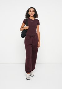 Burgundiškas sportinis kostiumas, sudarytas iš prigludusio T-shirt ir laisvų jogger tipo kelnių, abu pagaminti iš minkštos medžiagos, derinamas su lengvais sportbačiais ir juoda rankine.