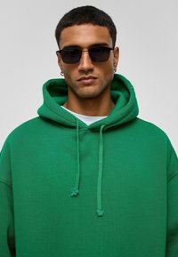 Groene hoodie met een zachte textuur en een trekkoordcapuchon, gedragen over een wit shirt. Accessoires zijn onder andere zwarte rechthoekige zonnebril.