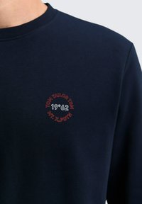 Námornícky modré tričko s kruhovým výstrihom a malým červeným a bielym textovým logom na hrudi s nápisom "TOM TAILOR TRM 19°62 MT. X. PDTN".