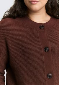 Bruine geribbelde cardigan met een ronde halslijn, voorzien van drie zwarte knopen aan de voorkant en een zachte, textuurachtige stof.