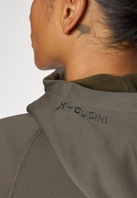 Houdini fleece hoodie in olijfgroen, met een zachte textuur, hoge kraag en geborduurd logo op de schouder. Zichtbare naaddetails.