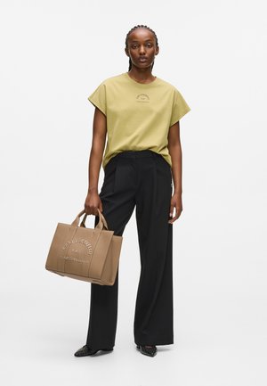 Olijfgroene oversized T-shirt, zwarte wijde broek, en tan handtas met textuur en reliëflogo, met daarbij puntige zwarte gestikte schoenen.