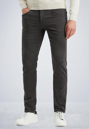 Mann, der dunkelgraue, schmal geschnittene Hose und weiße Sneaker trägt, mit einer Hand in der Tasche und der anderen seitlich am Körper.