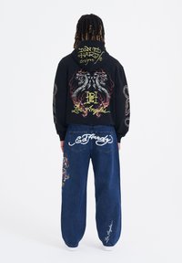 Felpa nera con un grafico colorato di un drago e testo sul retro, abbinata a jeans blu larghi con ricami e loghi.