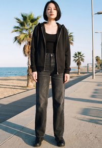 Fiatal nő rövid fekete hajjal, fekete kapucnis pulóvert, fekete trikót és sötét farmert visel, és egy napsütéses tengerparti sétányon áll, ahol pálmafák vannak.