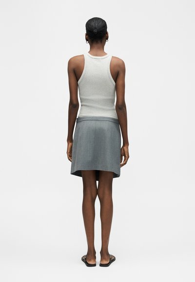 Γυναίκα με κοντά μαλλιά, φορώντας λευκό τοπ χωρίς μανίκια, γκρι φούστα και μαύρα σανδάλια, στέκεται πλάτη σε ένα απλό φόντο.