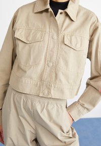Przycięta beżowa kurtka dżinsowa z zapięciem na guziki, dwiema kieszeniami piersiowymi i spiczastym kołnierzem, połączona z jasnobeżowymi spodniami na elastycznej talii.