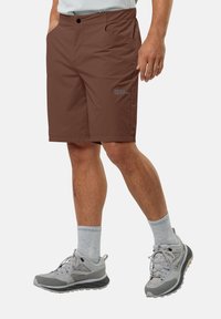 Shorts marron en tissu léger et respirant. Les caractéristiques incluent une poche avant, des détails de logo et une coupe décontractée. Portés avec des chaussures de sport grises.