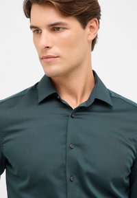 Camicia button-up verde scuro con un colletto strutturato, maniche lunghe e bottoni neri. Tessuto liscio con una sottile texture.