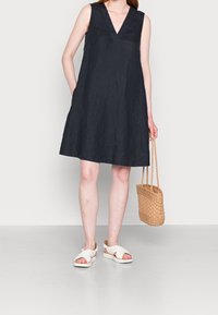 Robe sans manches en lin bleu marine avec un décolleté en V, coupe loose, poches latérales et un sac à main tissé beige. Le mannequin porte des sandales blanches.