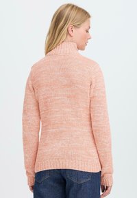 Kvinne med blondt hår iført en ferskenfarget strikket turtleneck genser og mørkeblå jeans, vist fra ryggen mot en ensfarget bakgrunn.