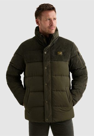 Homme portant une veste matelassée vert foncé avec un col montant et une poche zippée sur la manche gauche, debout, les mains dans les poches.