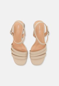Sandales à talons beiges avec une texture lisse, présentant trois brides superposées et une bride de cheville, avec une semelle intérieure de couleur claire.