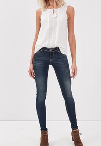 Mujer con blusa sin mangas blanca, jeans ajustados oscuros y botines de cordones marrones, de pie contra un fondo blanco liso.