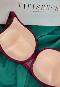 Gevormde bh met een naadloze nude cup, omrand met een bordeauxrode rand, met gaasdetails en verstelbare banden, rustend op groene stof.