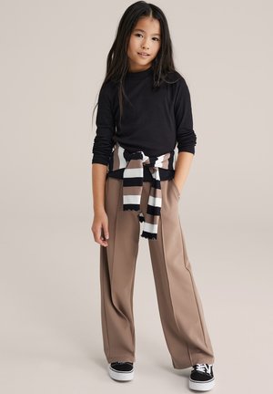 Junges Mädchen mit langen dunklen Haaren, das ein schwarzes langärmeliges Oberteil, beige weit geschnittene Hosen, schwarze Turnschuhe und einen gestreiften Pullover um die Taille gebunden trägt.