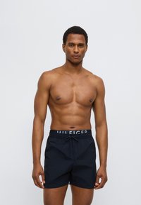 Μαύρα μαγιό σορτς με λάστιχο στη μέση και κορδόνι, καθώς και ελαστική ταινία με λογότυπο. Το ύφασμα είναι λείο και ελαφρύ, κατάλληλο για δραστηριότητες στο νερό.