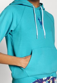 Turkos hoodie med en framficka, dragsko och en liten logotyp i en mörkare nyans. Tillverkad av bomull med en jämn textur.