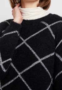 Zwarte gebreide trui met een pluizige textuur, voorzien van een wit ruitpatroon. De halslijn is rond en aansluitend.