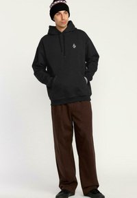 Sudadera negra con capucha, bolsillo frontal y logo, combinada con pantalones sueltos de color marrón oscuro y zapatos negros. Lleva un gorro de lana negro.