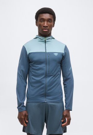 Młody mężczyzna stoi, ubrany w niebieską i jasnoniebieską bluzę na suwak z kapturem oraz pasujące spodenki, zwrócony twarzą do przodu na jednolitym tle.