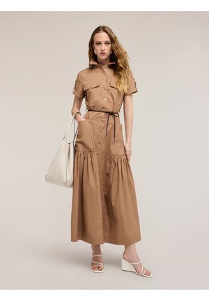 Donna con abito midi color sabbia con bottoni e cintura nera, che tiene una grande borsa bianca, indossa sandali bianchi con zeppa, in piedi su sfondo semplice.