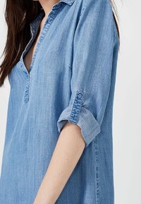 Světle modrá denimová košile s límcem a knoflíkovým manžetou. Tkanina má hladkou strukturu s jemnými barevnými variacemi.