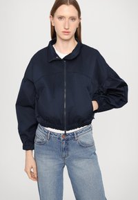 Marineblå cropped jakke med oppreist krage, elastiske mansjetter og glidelås foran. Kombinert med lyseblå høyt midjepants. Glatt tekstur.