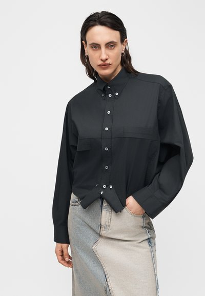 Άτομο που φοράει μαύρο πουκάμισο με κουμπιά, βιδωμένο άνισα σε φούστα από denim με δύο τόνους, με το ένα χέρι στην τσέπη, μπροστά σε απλό φόντο.