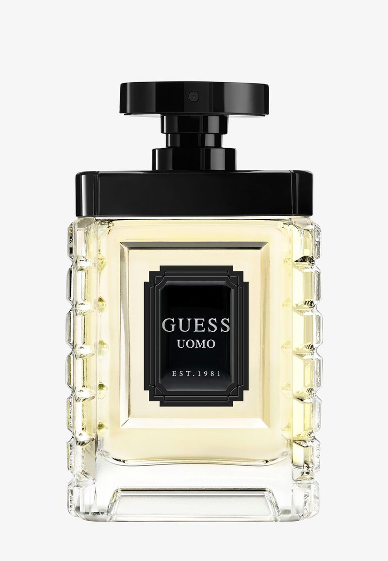 Gjennomsiktig glassparfymeflaske med en teksturert rektangulær form, svart lokk og etikett med teksten "GUESS UOMO EST. 1981" i hvit skrift.