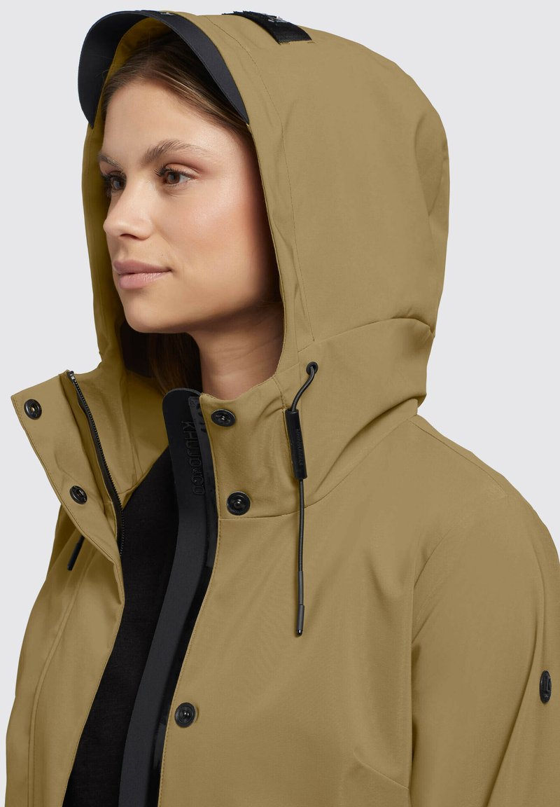 Veste khaki à capuche avec une doublure intérieure noire, cordon de serrage ajustable et boutons-pression. Tissu lisse avec une finition mate et matériel dissimulé.