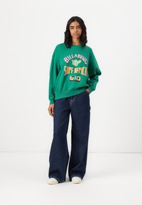 Felpa verde a collo rotondo con vestibilità oversize, caratterizzata da una grafica a tema surf in giallo e rosso. Abbinata a jeans scuri a gamba larga e sneakers.