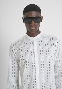Witte, langarmige shirt met verticale uitsnijdingen, knoopsluiting en een hoge kraag. Draagt oversized zwarte zonnebril.