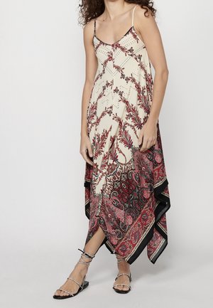 Kobieta ubrana w bezrękawnikową kremową sukienkę z czerwonymi kwiatowymi i paisley wzorami oraz czarne, płaskie sandały na paski, stojąca na jednolitym tle.