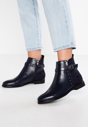 Bottes en cuir bleu marine montant à la cheville avec une fermeture éclair latérale, une sangle à boucle et des détails cloutés, portées avec un jean court bleu clair.