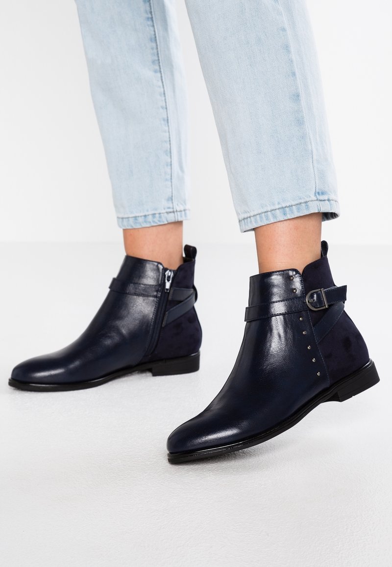 Bottes en cuir bleu marine montant à la cheville avec une fermeture éclair latérale, une sangle à boucle et des détails cloutés, portées avec un jean court bleu clair.
