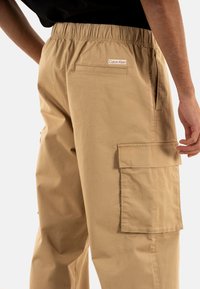Beige cargobroek met een elastische tailleband, zakken aan de zijkant en een klepzak op de dij. Heeft een Calvin Klein-label op de achterkant.