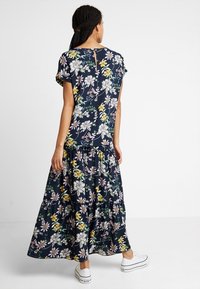 Femme avec des cheveux tressés portant un haut à fleurs navy à manches courtes et une jupe longue assortie, associée à des baskets blanches, se tenant de dos.