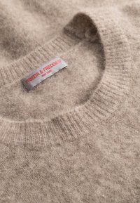 Beige wollen trui met een geribde ronde hals, voorzien van een grijs label met rode tekst "FRIEDA & FREDDIES® NEW YORK." Zachte, fuzzy textuur.