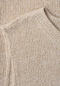 Pull beige en maille avec texture visible et col ainsi que bords de manches côtelés, posé à plat sur un fond en tissu assorti.