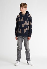 Sudadera marina con patrones geométricos beige, bolsillo frontal y capucha con cordón, combinada con jeans grises y zapatillas blancas.