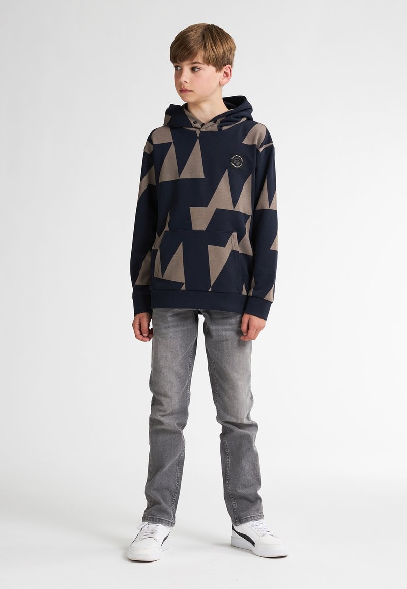 Sudadera marina con patrones geométricos beige, bolsillo frontal y capucha con cordón, combinada con jeans grises y zapatillas blancas.