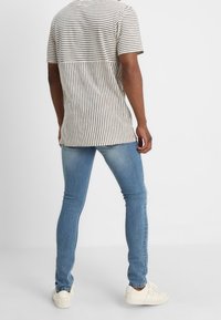 Camisa de manga corta a rayas en beige y marrón oscuro, combinada con jeans ajustados de color azul claro y zapatillas blancas, vista desde la parte posterior.