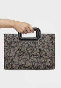 Dłoń trzyma prostokątną torebkę z czarnym abstrakcyjnym wzorem oraz czarnym prostokątnym uchwytem na jednolitym białym tle.