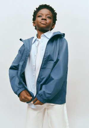 Jongen in een blauwe capuchonjas en een wit overhemd die de rits van de jas dichtdoet tegen een effen achtergrond.