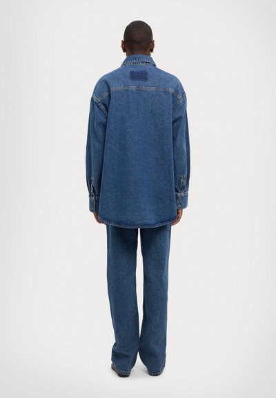 Denimjakke og bukser i mellomblått. Jakken har spiss krage, knappemansjetter og en avslappet passform. Myk tekstur, uten synlige mønstre.