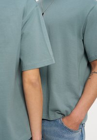 Δύο άτομα με χλωμό πράσινο κοντομάνικο πουκάμισο, ο ένας φοράει κολιέ, στεκόμενοι κοντά, με το ένα χέρι στην τσέπη του τζιν και με ένα ορατό τατουάζ στο χέρι.