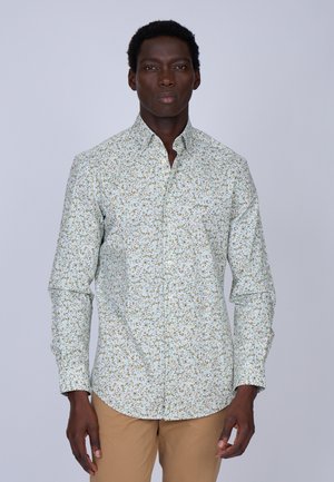 Chemise blanche à manches longues avec un motif floral vert et jaune, dotée d'un col pointu et d'une fermeture à boutons, associée à un pantalon beige.